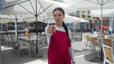 Kırmızı önlüklü genç bir kadın caddede şemsiyeli ve sandalyeli bir kafenin terasında el kol hareketi yapıyor.