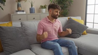 Gri bir koltukta oturan, pembe tişörtlü bir adam modern bir oturma odasında hayal kırıklığı içinde, dolapta bitkiler ve kitaplarla oturuyor.