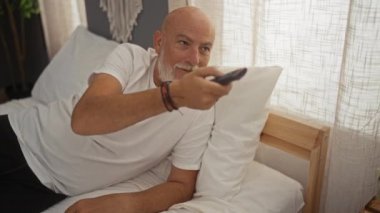 Bir adam evinde, yatak odasında uzanıyor. Uzaktan kumandayla rahat bir ev ortamında, gri sakallı, mutlu ve mutlu görünüyor..