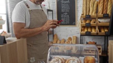 Fırındaki yaşlı adam, ekmek rafları ve tatlılarla çevrili telefonla notlar alıyor.