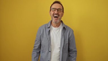 İspanyol orta yaşlı gözlüklü bir adam çılgın bir ifadeyle ayakta duruyor hayal kırıklığı içinde bağırıyor, kollarını kaldırıyor, izole edilmiş sarı arka planda çılgın bir öfke patlaması yaşıyor.