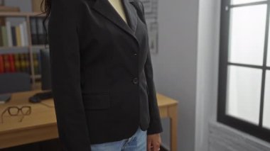 Ofiste kolları çapraz duran, siyah ceketli, kumral saçlı, tahta masada gözlüklü, kitaplık arka planda, iş yerinde, kapalı alanda duran bir kadın.