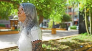 Gülümseyen bir kadın, yemyeşil bir arka planı ve şehir binaları olan bir şehir parkında duruyor gri saçlarını ve dövmeli kollarını gösteriyor..