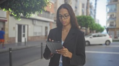 Şehir binaları ve arka planda arabası olan şehir sokaklarında tablet kullanan gözlüklü genç bir kadın modern teknoloji ve profesyonel yaşam tarzını gözler önüne seriyor.