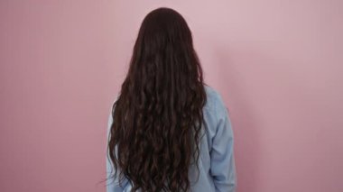 Pembe bir duvarın önünde gülümseyen, açık mavi gömlekli, uzun esmer saçlı bir kadın..