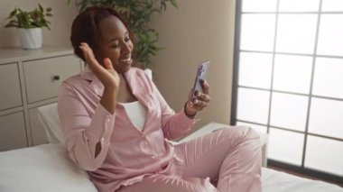 Pembe pijamalı genç bir kadın rahat yatak odasında akıllı telefon kullanıyor.