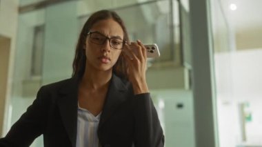 Modern ofiste akıllı telefondan sesli mesaj dinleyen gözlüklü ve takım elbiseli bir kadın.