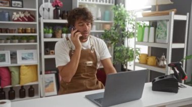 Önlüklü genç adam ev dekorasyonu mağazasında dizüstü bilgisayar kullanırken telefonla konuşuyor. Arka planda çeşitli dekorasyonlar ve renkli yastıklar var.