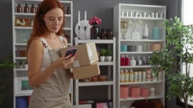 Bir ev dekorasyonu dükkanında elinde paketler olan ve etrafı kapalı bitkiler ve dekoratif malzemelerle çevrili akıllı telefon kullanan genç bir kadın.