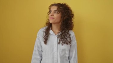 Tişörtlü İspanyol genç bir kadın, yanlamasına ayakta duruyor, gülümsüyor ve izole edilmiş sarı arka plan üzerinde düşünüyor.