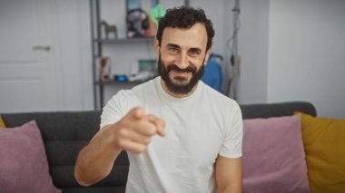 Modern bir oturma odasında günlük giysiler içinde yakışıklı, sakallı ve gülümseyen adam.