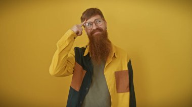 Bearded young man with glasses gesturing to his head, isolated on a yellow background, portraying thinking or having an idea. clipart