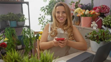 Canlı çiçekçide bilgisayarın başında akıllı telefon kullanan gülümseyen kadın.