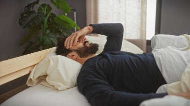 Yatakta yatan sakallı bir adam evde yatak odası düzeninde gergin ya da hasta görünür..