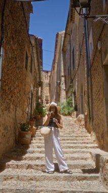 Genç bir kadın Fornalutx, Mallorca, İspanya 'nın büyüleyici sokaklarında güneşli bir günün tadını çıkarıyor. Yazlık giysiler ve şapka giyiyor. Geleneksel taş binalar ve renkli bitkilerle kaplı..
