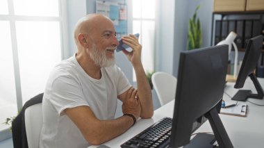 Ofiste gri saçlı ve sakallı bir adam telefonda konuşuyor. Kapalı alanda bilgisayarla çalışıyor. Doğal ışıklandırma ve günlük kıyafetlerle çalışıyor.