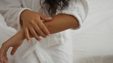 Beyaz bornozlu bir kadın kapalı bir yatakta oturuyor ve rahat bir yatak odasında koluna hafifçe dokunuyor. Huzurlu ve rahat bir an sergiliyor..