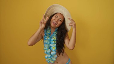 Sarı arka planda poz veren güzel bir genç kadın şapka ve Hawaii zambağı takıyor, neşeli bir barış işareti ve gülümseme ile, yaz titreşimleri yayıyor ve neşeli bir enerji yayıyor.