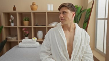 Spa 'da bornoz giyen genç bir adam huzur dolu bir salonun içinde rahatlama deneyimi yaşıyor. Etrafı raflarla çevrili ve rahatlatıcı bir ortam.
