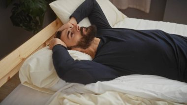 Sakallı adam modern bir yatak odasında dinleniyor, sakinlik ve rahatlık yayıyor..
