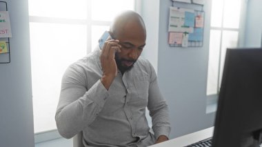 Yakışıklı, Afro-Amerikalı, sakallı, kel bir adam. Ofiste oturuyor, cep telefonuyla konuşuyor ve bilgisayar üzerinde çalışıyor..