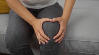 Kapalı bir yerde oturan genç bir kadın dizini kavrıyor, ev içi arka planda acı ya da rahatsızlık tasvir ediyor..