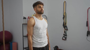 Yakışıklı, sakallı, spor salonunda beyaz atletli, spor aletleriyle çevrili genç bir adam.