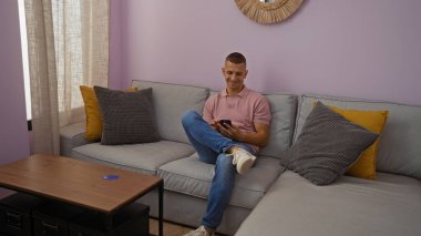 Genç adam rahat bir oturma odasında, etrafında yastıklar ve iyi aydınlatılmış bir evin içindeki kahve masası olan akıllı bir telefon kullanarak koltukta oturuyor.