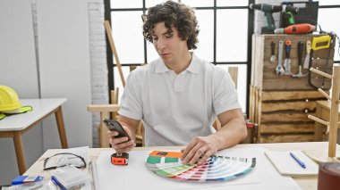 Hispanic man examines color swatches in a well-equipped carpentry workshop while holding a smartphone. clipart