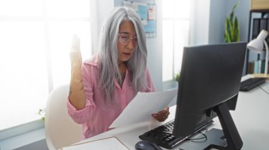 Ofiste elinde evrak tutan bir kadın, olgun ve gri saçlı, kapalı bir masada çalışan, odaklanmış ve profesyonel görünen..