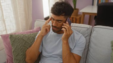 Genç İspanyol adam telefonda konuşuyor. Oturma odasında kanepede oturuyor. Düşünceli bir ifadeyle.