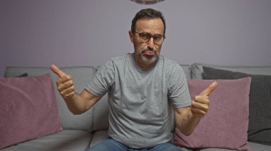 Yakışıklı, orta yaşlı, suratını buruşturan, parmakları havada, evdeki oturma odasındaki kanepede oturan adam.