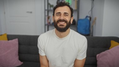 Sakallı, beyaz tişörtlü, orta yaşlı neşeli bir adam, iyi aydınlatılmış bir oturma odasında gri bir koltukta oturuyor..