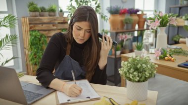 İspanyol kadın çiçekçi dükkanında çiçek ve çiçeklerle çevrili notlar yazarken sesli mesaj dinliyor..