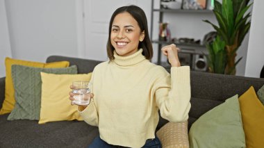 Sıcak bir oturma odasında renkli yastıkları olan bir koltukta oturan, elinde bir bardak su tutan gülümseyen İspanyol kadın..