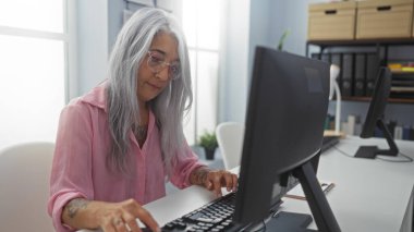 Kafkasyalı, gri saçlı, yaşlı bir kadın. Kapalı bir ofiste bilgisayar başında çalışıyor. Arkaplanda raflar ve masa var..