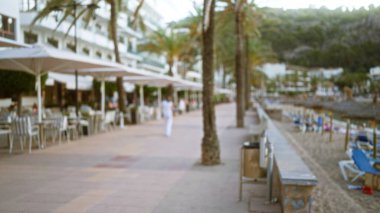 Mallorca 'daki dış mekanın bulanık arkaplanı, palmiye ağaçları, açık hava oturma alanları ve uzakta duran beyaz elbiseli birini gösteriyor..