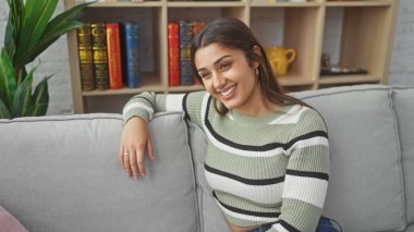 Neşeli, genç, İspanyol bir kadın. Gri bir kanepede dinleniyor. Güzellik ve rahatlık saçıyor..