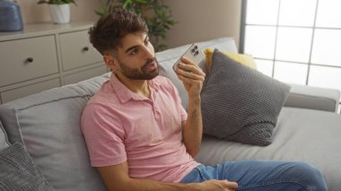 Kanepede rahat bir oturma odasında oturan, akıllı telefondan konuşan, sakallı genç İspanyol bir adam..