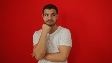 Genç İspanyol adam izole edilmiş kırmızı bir arka plana karşı düşünceli bir şekilde poz veriyor, beyaz bir tişört giyiyor, çekici bir yetişkin erkek görünümünü gündelik bir ortamda sergiliyor..