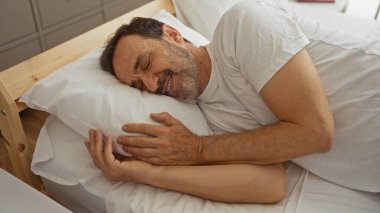 Yakışıklı, orta yaşlı bir adam. Yatağında uzanmış, rahatça dinlenirken sıcak bir şekilde gülümsüyor.