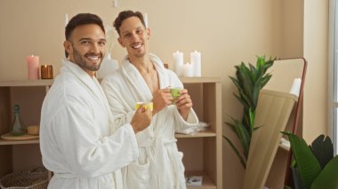 Bornozlu eşcinsel çift, rahat bir spa ortamında mumlar ve bitkiler eşliğinde, sevgi, birliktelik ve refahı ön plana çıkaran kupalarla birlikte rahatlama zamanının tadını çıkarıyor..