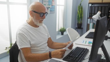 Modern bir ofiste sakallı kıdemli bir adam masasında doğal ışık altında belgeleri inceliyor.