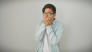 Gözlüklü genç Asyalı bir adam beyaz bir arka plana karşı ağzını kapatır, sürpriz veya gizlilik iletir..