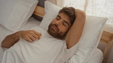 Sakallı genç adam rahat bir yatak odasında uyuyor. Yumuşak yastıklar ve pencereden içeri giren güneş ışığıyla huzurlu bir ortam sergiliyor..
