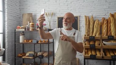 Fırındaki gri saçlı adam içeride neşeyle gülümserken çeşitli ekmekleri gösteren video çağrısı yapıyor.