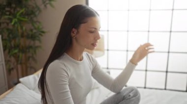 Yatak odasında, el ele tutuşan, sıkıntılı görünen, gri elbiseli, pencereli ve arka planda bitkiler olan bir kadın.