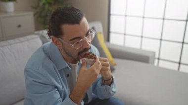 Evdeki rahat oturma odasında gözlüklü ve kek yiyen genç adam rahat bir yaşam tarzı sergiliyor.