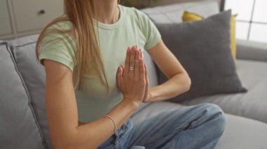 Genç, güzel, sarışın bir kadın huzurlu bir oturma odasında meditasyon yapıyor, gözleri kapalı, sakin bir evde rahat bir koltukta oturuyor..