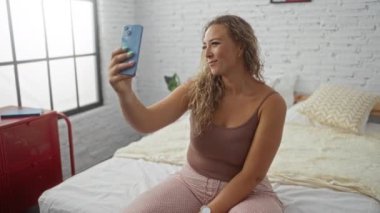 Yatak odasında yatakta oturan bir kadın akıllı telefonuyla selfie çekiyor. Gülümsüyor ve mutlu görünüyor.